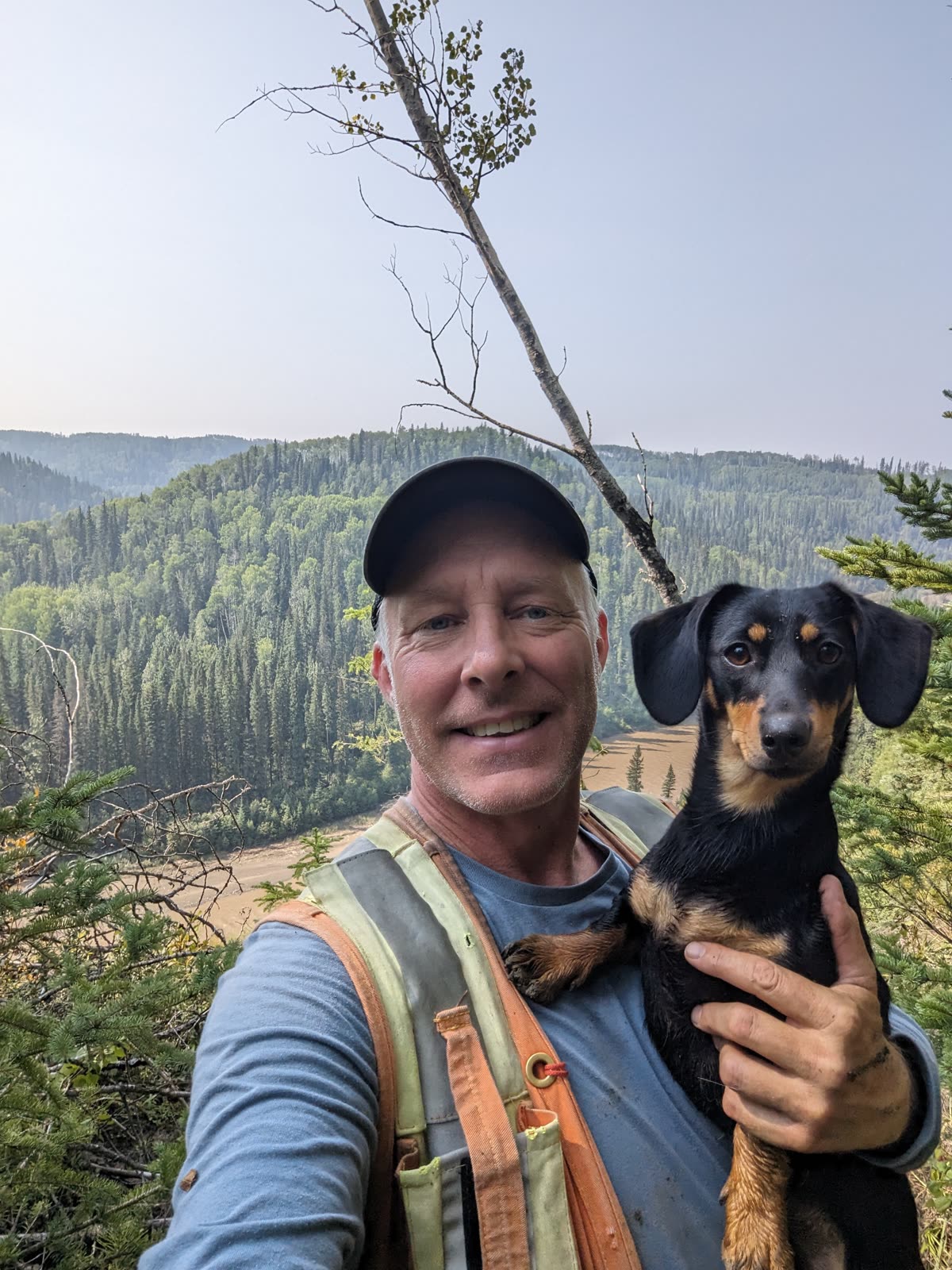Derek in a cap and hiking vest holding Lola while standing on a ridge above a forested valley